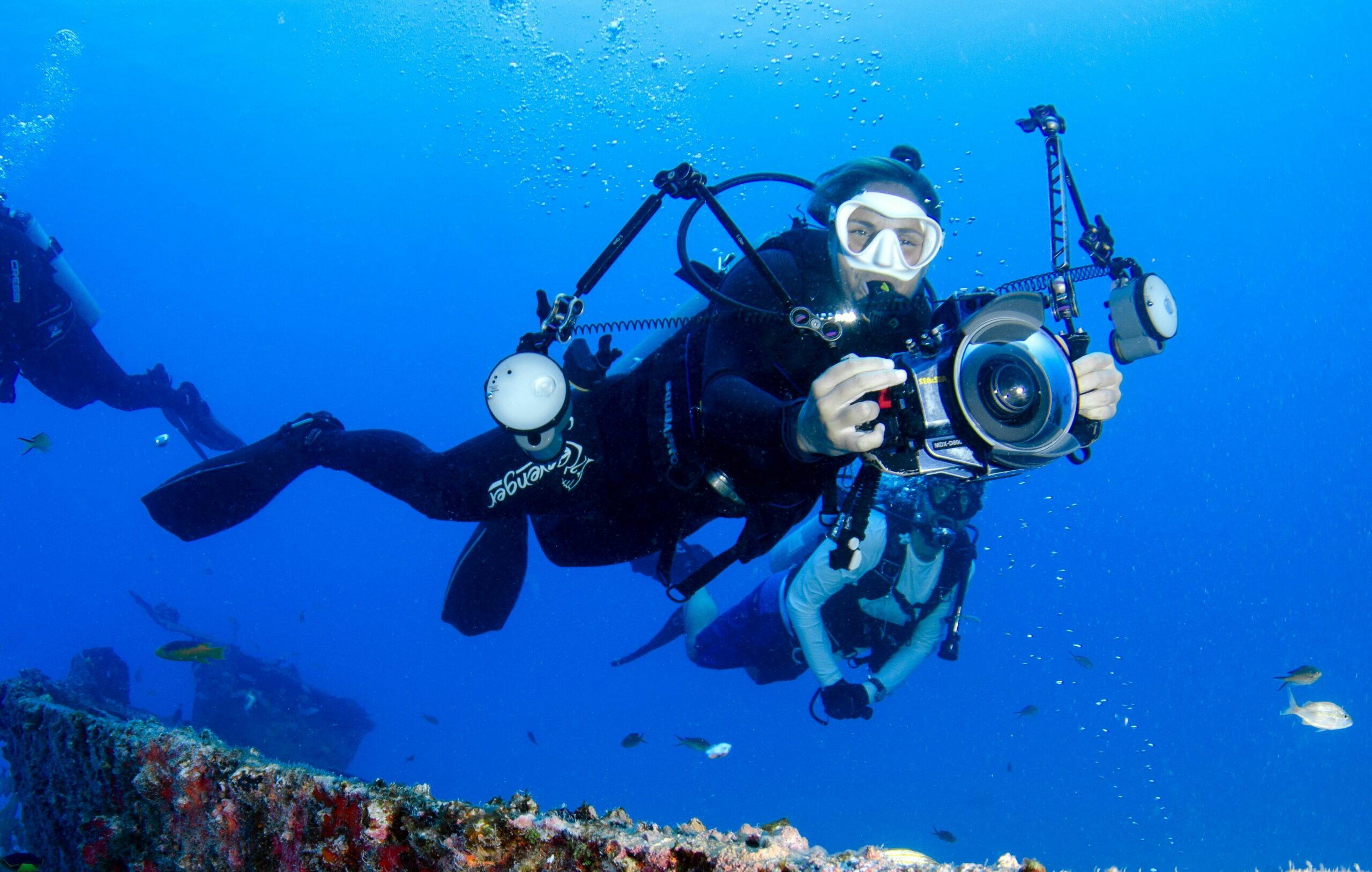 Two scuba divers capturing the vibrant marine life near an underwater wreck in crystal clear waters.